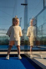 Blond young boy with topknot standing on blue tennis court near glass wall, holding a green ball in one hand, smiling with reflection visible in the glass