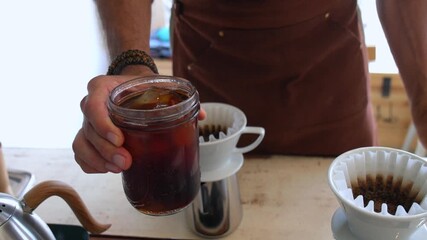 Professional male barista in a coffee shop adding ice cubes to a freshly brewed cold brew coffee in a glass jar, then stirring it with a straw before serving the refreshing summer beverage - Powered by Adobe