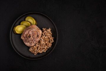 Turkey steak with buckwheat porridge and pickled cucumber on a round black plate against a black background. Photo of prepared food with copy space. Natural healthy eating concept. Top view
