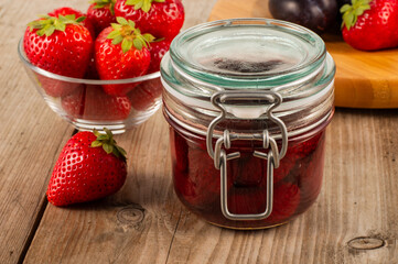 Glass jar of strawberry jam placed on a wooden table with fresh strawberries in the background, creating a natural fruit and homemade food concept ideal for culinary and recipe uses