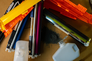 Colorful printer cartridges and wasted toner bottles scattered on a cardboard surface with spilled black powder.