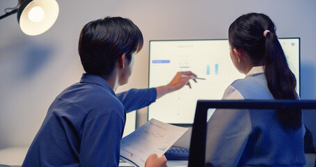 Two young Asian business colleagues are analyzing financial data and charts on a desktop computer screen in a modern office late at night.