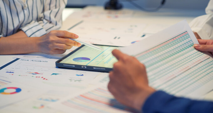 Close-up business team analyzing financial charts and data together, using a tablet and printed reports during a meeting. Hands pointing at graphs while discussing strategy and performance metrics.