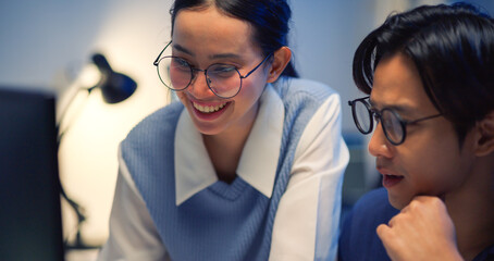 Close-up portrait of two young Asian business colleagues smiling and looking satisfied while analyzing data late at night. Conveys happiness, success, and positive teamwork.