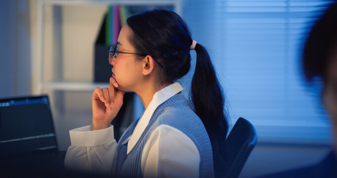 A young Asian woman wearing glasses sits at her desk, concentrating intensely while working on a laptop late at night in a cool-toned office. Conveys dedication and overtime work.