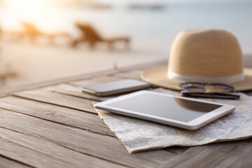 Straw hat, tablet, smartphone, map, sunglasses laid on wooden table; peaceful beach getaway awaits.