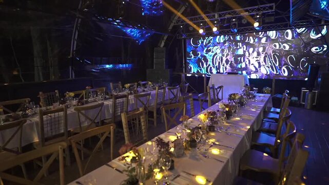 Wide, empty event hall features long banquet settings, detailed table decor, staged blue and gold lighting, and modern AV setup, ready for a formal celebration.