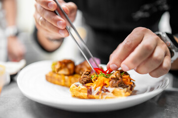 A chef in a black uniform uses precision tweezers to garnish a gourmet dish, which appears to be braised meat and shredded vegetables served on a waffle, in a professional kitchen setting.