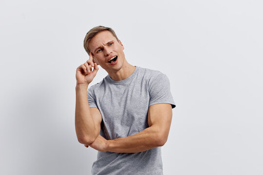 Young confused caucasian man wearing gray t-shirt thinking with hand near ear isolated on light background puzzled expression head tilt gesture. Mental health concept