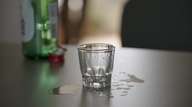 Empty Soju Shot Glass on Table with Spilled Alcohol – Korean Drink Scene. A silhouette of a person pacing in the background - Powered by Adobe