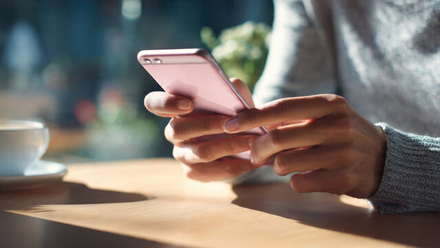 Close-up of hands holding a pink smartphone at a cafe table with a coffee cup, illuminated by sunlight, representing modern digital interaction. - Powered by Adobe