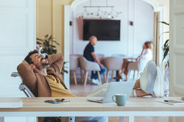 Relaxed Businessman At Modern Office Desk With Laptop And Colleagues In Background