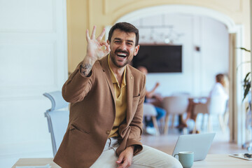 Smiling Man in Brown Blazer Waving OK Gesture in Modern Office Setting for Business