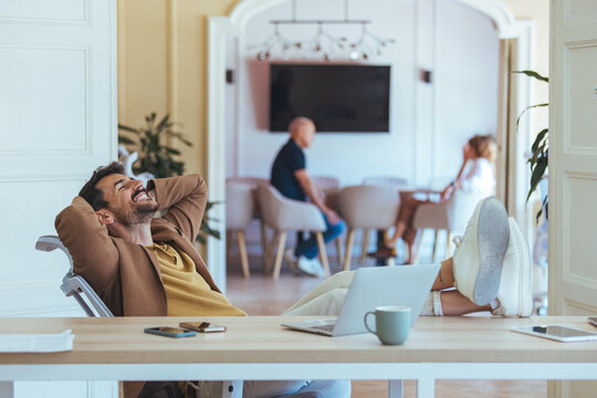 Relaxed Businessman At Modern Office Desk With Laptop And Colleagues In Background