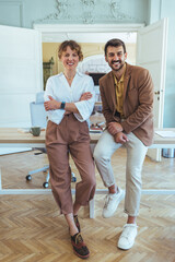 Two Professionals in Casual Business Attire Smiling While Leaning on a Desk in a Bright Office