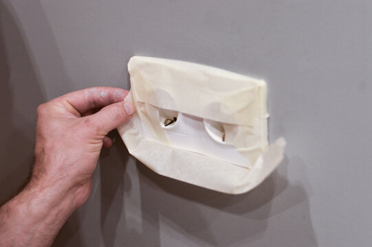 Focused on a pair of hands, this image demonstrates the practical step of masking an electrical outlet with durable tape to safeguard it from accidental paint damage during home repairs.