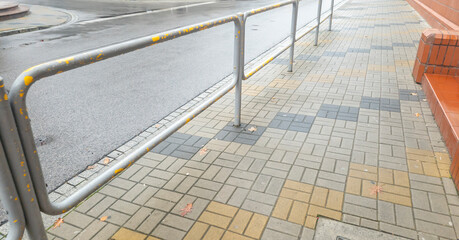A view of a public sidewalk featuring a metallic railing alongside a pattern of concrete tiles in warm colors. The scene is set on a rainy day, reflecting urban elements.