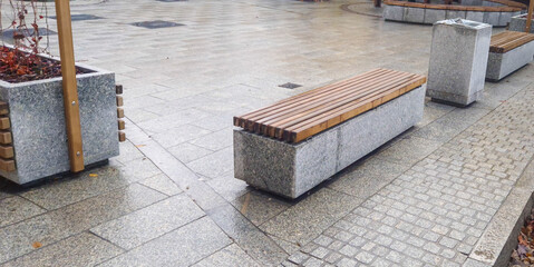 A modern urban park with sleek stone benches and wooden accents, providing a public resting area....