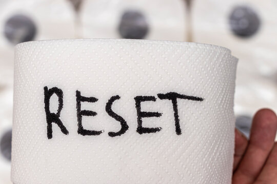 Handwritten RESET on a roll of toilet paper conveys a message of renewal and fresh beginnings, set against a blurred background of additional toilet paper rolls.