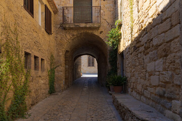 Narrow cobblestone street in Peratallada’s old town, passing under stone archways with rustic walls and climbing plants.