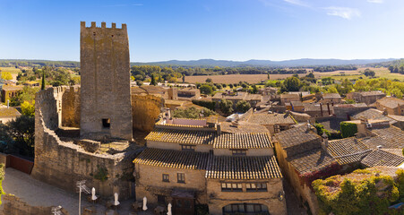Panoramic view of Peratallada with the medieval castle tower rising over tiled rooftops, surrounded by countryside under a clear blue sky.