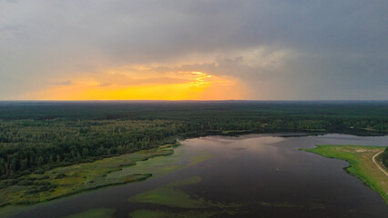 Aerial view of a serene landscape at sunset, featuring a tranquil lake surrounded by lush greenery and forest under a moody sky, creating a peaceful atmosphere.