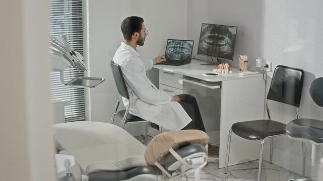 Male dental professional sitting at modern clinic workstation reviewing patient teeth X-ray on two displays for proper diagnostic assessment