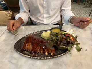Closeup shot of a male holding a knife getting ready to eat ribs pork steak with sauce and fresh vegetables on the table and green salad in a restaurant
