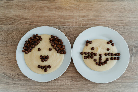 Two plates of porridge decorated with chocolate cereal pieces forming cute faces on a wooden table. - Powered by Adobe