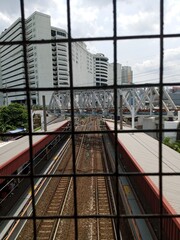 A view down onto train tracks and station platforms, framed by a dark wire mesh fence, with a steel truss bridge and tall white buildings visible in the background.