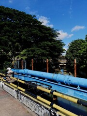 A bright outdoor shot featuring a large, blue industrial pipe or mechanism situated over a waterway, bordered by a yellow and black barrier and lush green trees against a blue sky.