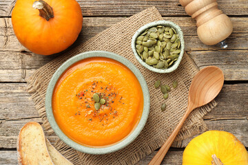 Tasty pumpkin cream soup with seeds in bowl, roasted bread and vegetables on wooden table, flat lay