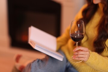 Woman with red wine reading book near fireplace at home, closeup