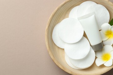 Clean cotton pads, plumeria flowers and tube of cosmetic product in wooden bowl on beige background, top view. Space for text