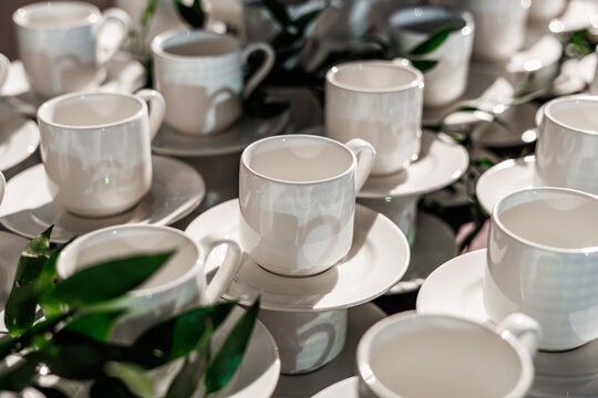 A neat arrangement of stacked white porcelain coffee cups and saucers on a table, accented with fresh green foliage. Perfect for catering or break service themes