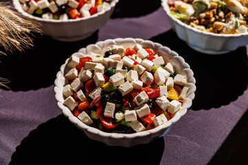 A vibrant Greek salad with diced cucumbers, tomatoes, olives, and bell peppers, topped with cubes of feta cheese in a scalloped white bowl on a dark tablecloth