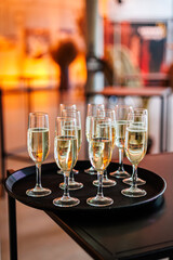 A black tray holding multiple champagne flutes filled with sparkling wine, set on a dark table with a warm, blurred background. Ideal for event or hospitality themes