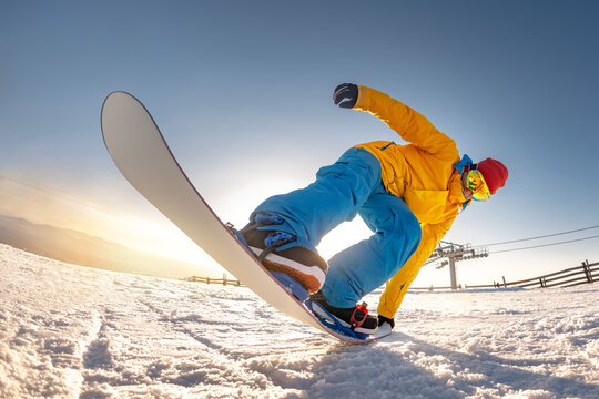 Young snowboarder in bright yellow and blue clothes makes trick "Tail press" at ski resort mountain top at sunset time