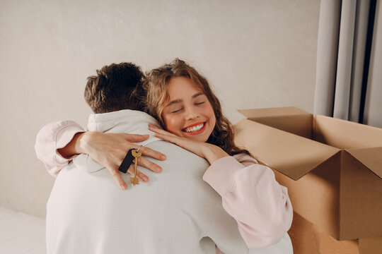 Happy young family couple man and woman hugging and holding keys from new home. Moving with cardboard boxes to new estate apartment.