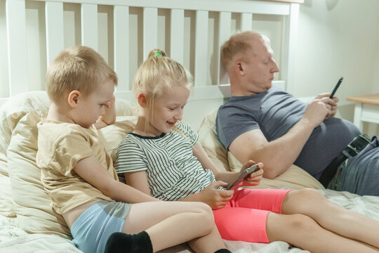 Joyful siblings and distracted father using phones and tablet on cozy bedroom bed. Family screen time in modern home interior