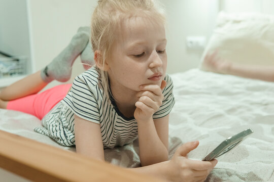 Young blonde girl lying on bed thoughtfully viewing tablet screen. Curious child pondering digital content in cozy bedroom with soft lighting