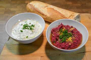 Two healthy dips, chickpea hummus with beetroot and vegan yogurt with cucumber, garlic and herbs, white bowls and bread on a rustic wooden board, selected focus