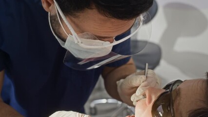 Male dentist wearing face mask and shield carefully examining patient teeth with dental tools while patient sitting on treatment chair in bright modern dental clinic