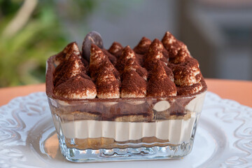 A glass dish of tiramisu showcases creamy layers, cocoa-dusted mascarpone, and a chocolate garnish on a white plate.