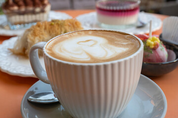 A white ceramic cup of cappuccino with heart-shaped latte art is surrounded by assorted desserts on an orange café table.