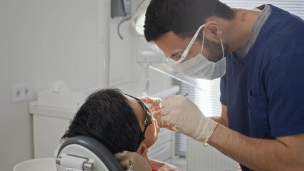 Professional Middle Eastern male dentist in uniform using dental tools while providing professional dental treatment to patient seated in treatment chair in clean modern dental clinic - Powered by Adobe