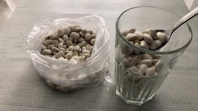 Kitchen Task: A close-up of a person using a tablespoon to transfer dry white beans from a faceted glass cup into a clear plastic bag for storage or portioning