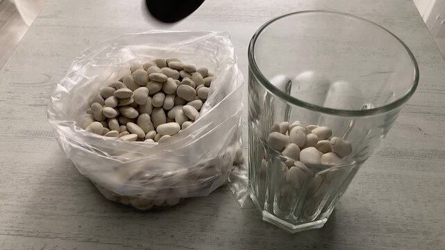 Kitchen Task: A close-up of a person using a tablespoon to transfer dry white beans from a faceted glass cup into a clear plastic bag for storage or portioning.