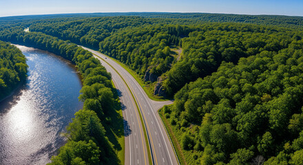 Aerial panoramic drone view of a riverside highway cutting through lush green forest with bright sunlight reflecting off the calm river beside the road