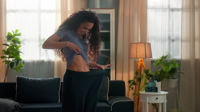 Excited Hispanic fit slim woman happy checking waist slimming good shape sport result weight lost sportswoman female fitness girl wearing old big jeans enjoying abs dieting healthy lifestyle at home